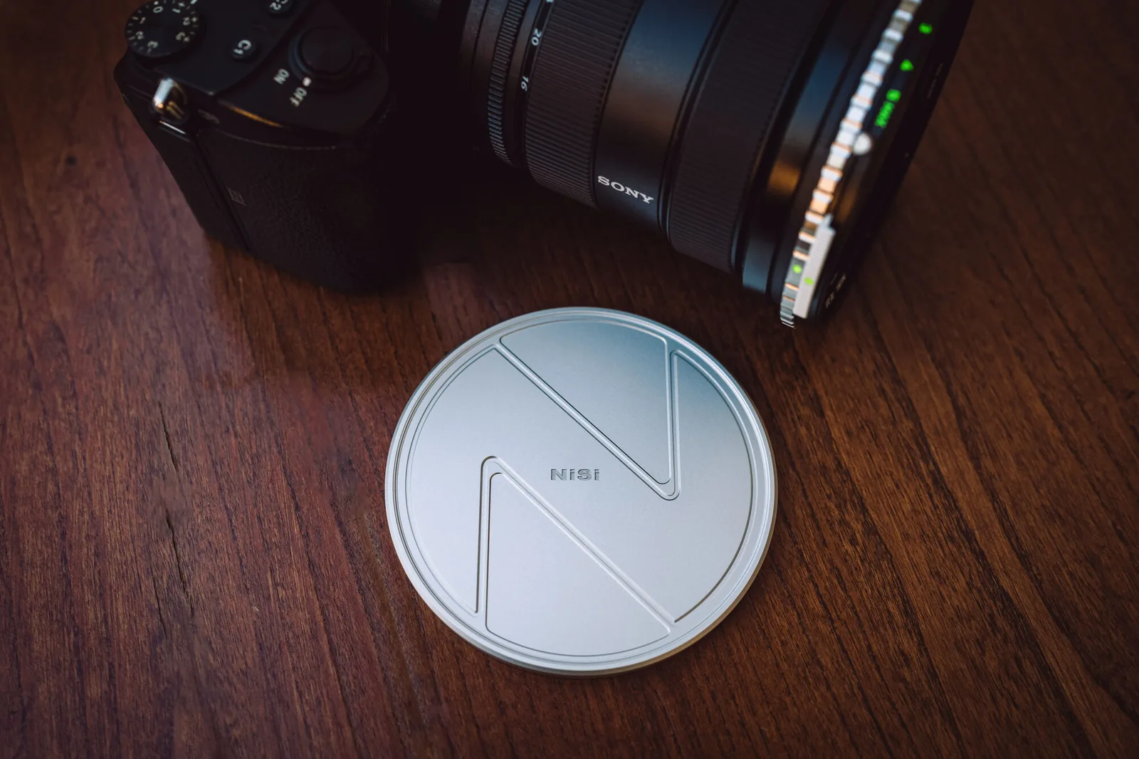 The silver machined aluminum NiSi lens cap on a wooden table beside a camera fitted with the filter