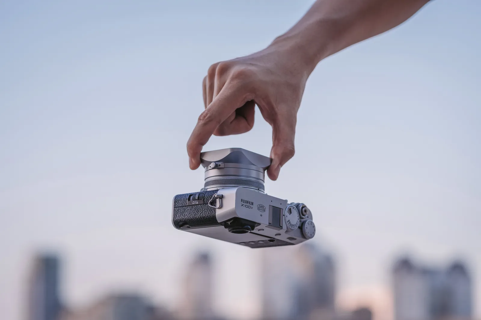 A silver Fujifilm X100V hanging from two fingers by its square NiSi lens hood alone, above an out-of-focus city skyline at dusk