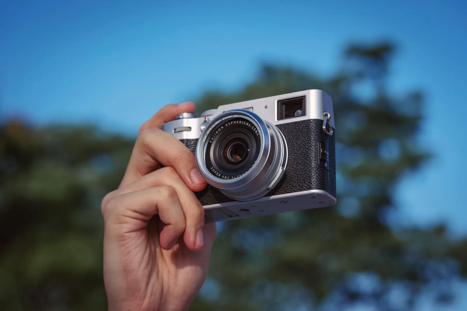 A silver Fujifilm X100 held up in one hand against a blue sky and out-of-focus trees, the NiSi filter fitted on the front of the lens