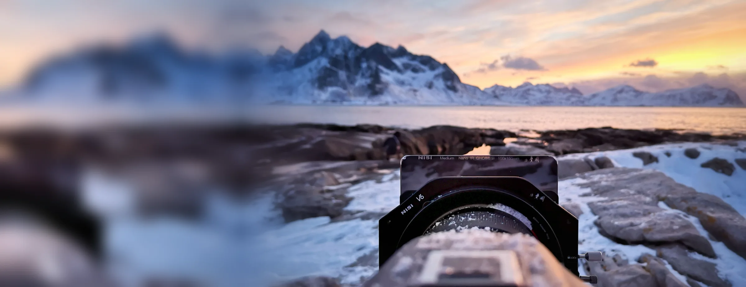 A NiSi 100mm filter holder with a graduated ND filter on a camera, set up low on snowy rocks facing a mountain range at dusk